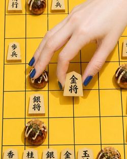 Jeu de shogi avec pièces en bois et takoyaki, main féminine déplaçant une pièce sur un plateau jaune.