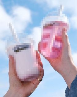 Dos manos sostienen vasos de bubble tea en un cielo azul con algunas nubes blancas visibles de fondo.