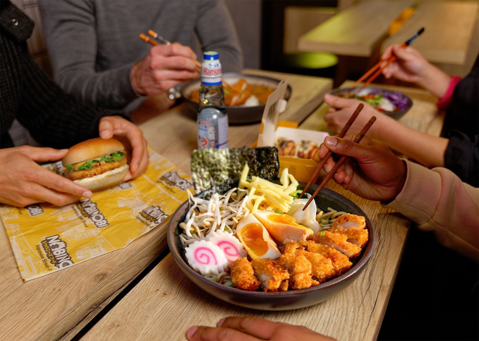 Plat de ramen avec œufs, morceaux de poulet frit et légumes, aux côtés d'un burger et d'une bouteille sur une table en bois.