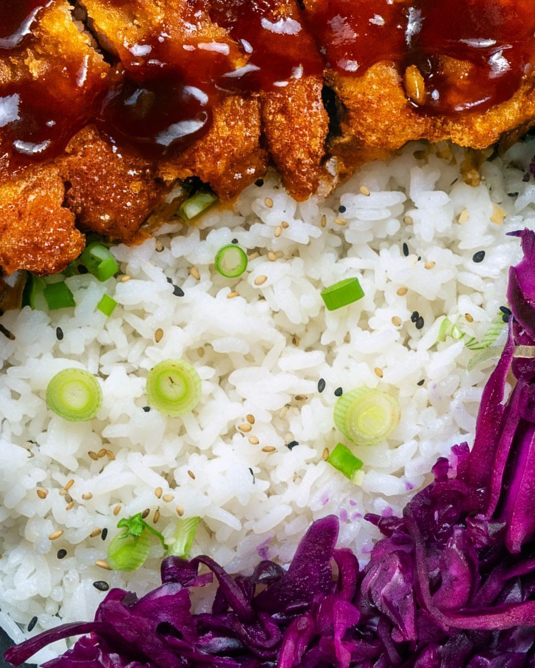 Close-up of crispy fried meat with sauce served alongside white rice garnished with green onions and purple cabbage.