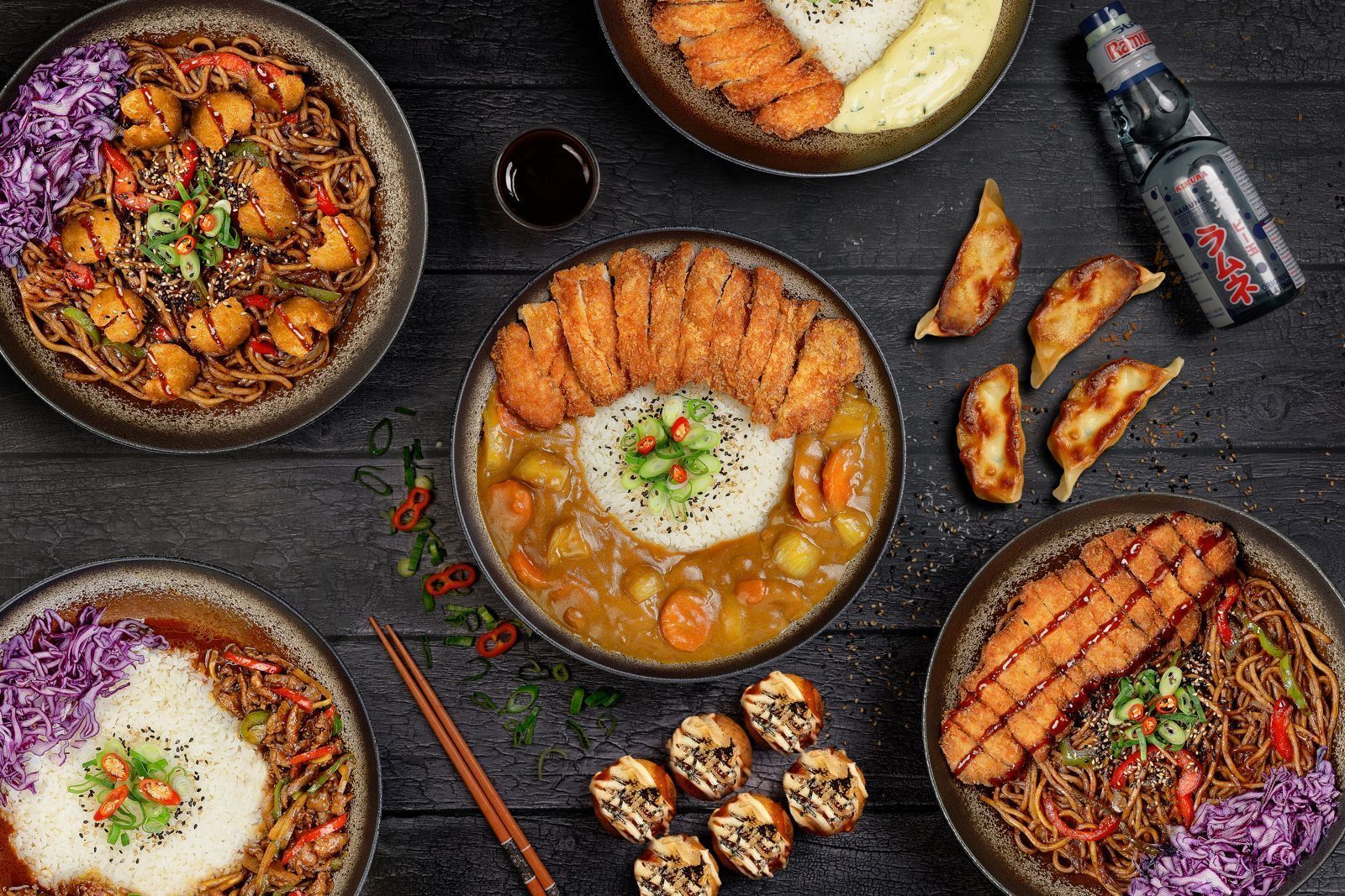 Platos variados de comida asiática con arroz, fideos, empanadillas y una botella de bebida sobre mesa oscura