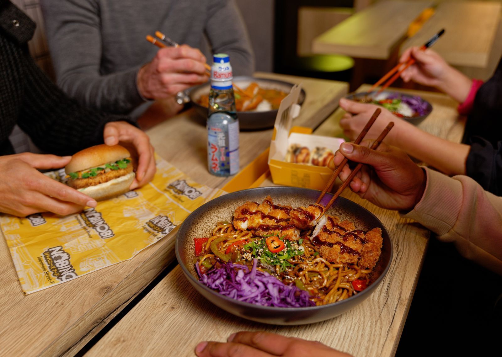 Plat de nouilles asiatiques avec poulet pané, chou rouge et légumes, accompagné d'un burger et boissons sur table en bois.