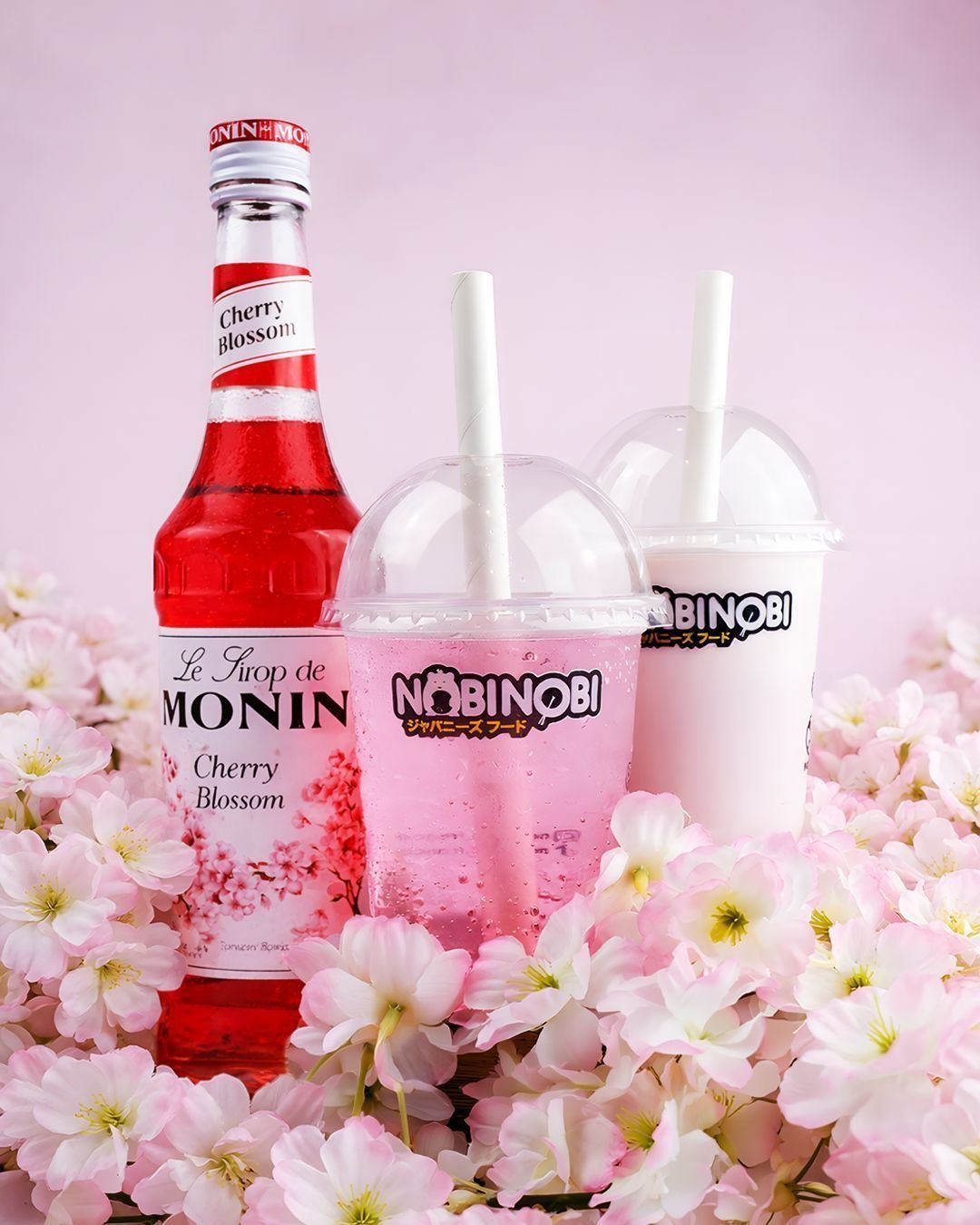 Two bubble tea drinks with Nobi Nobi logo surrounded by cherry blossom flowers and a bottle of cherry blossom syrup