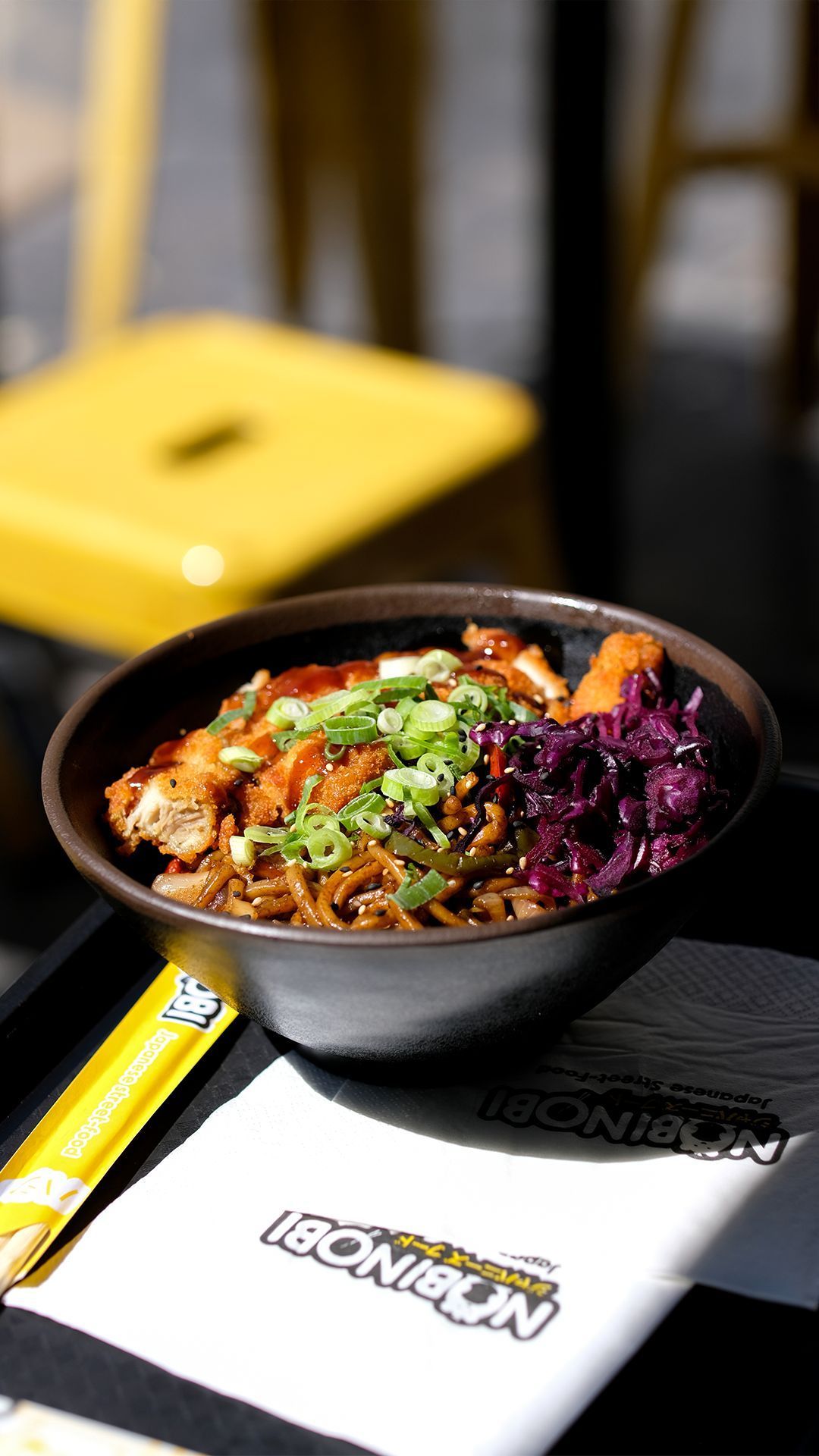 Tazón con fideos, pollo y repollo morado servido en el restaurante Nobi Nobi Bayonne con utensilios y servilleta visibles.