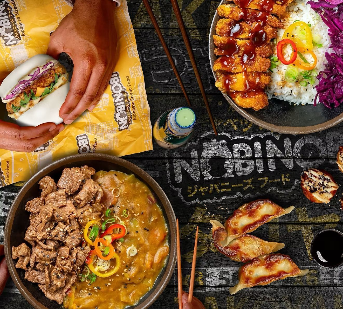 Asian restaurant dishes including ramen, bao burger, gyoza, and rice bowl with fried chicken on a table with chopsticks.