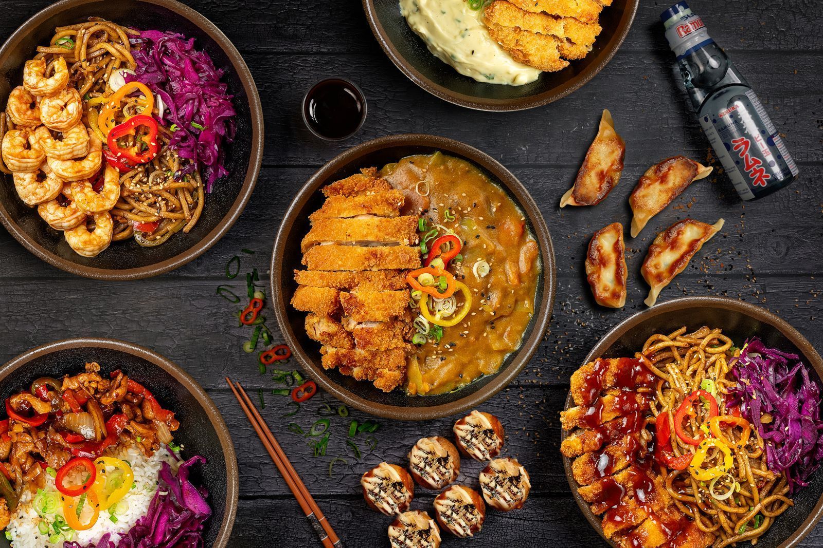 Plats asiatiques variés avec nouilles, riz, crevettes, katsu, dumplings et une boisson japonaise sur table noire.