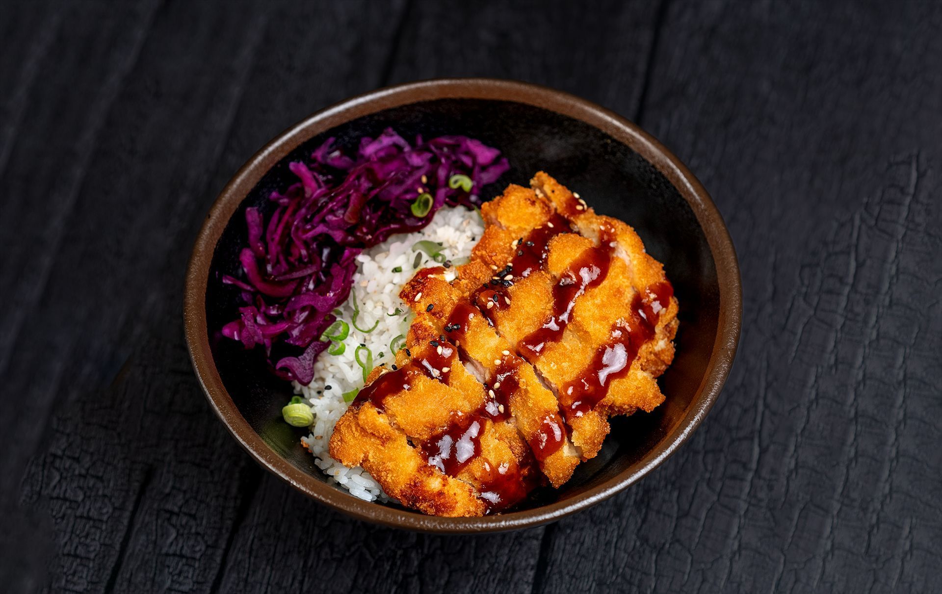 Bol con arroz, pollo empanizado con salsa y col morada en un fondo negro, comida típica asiática