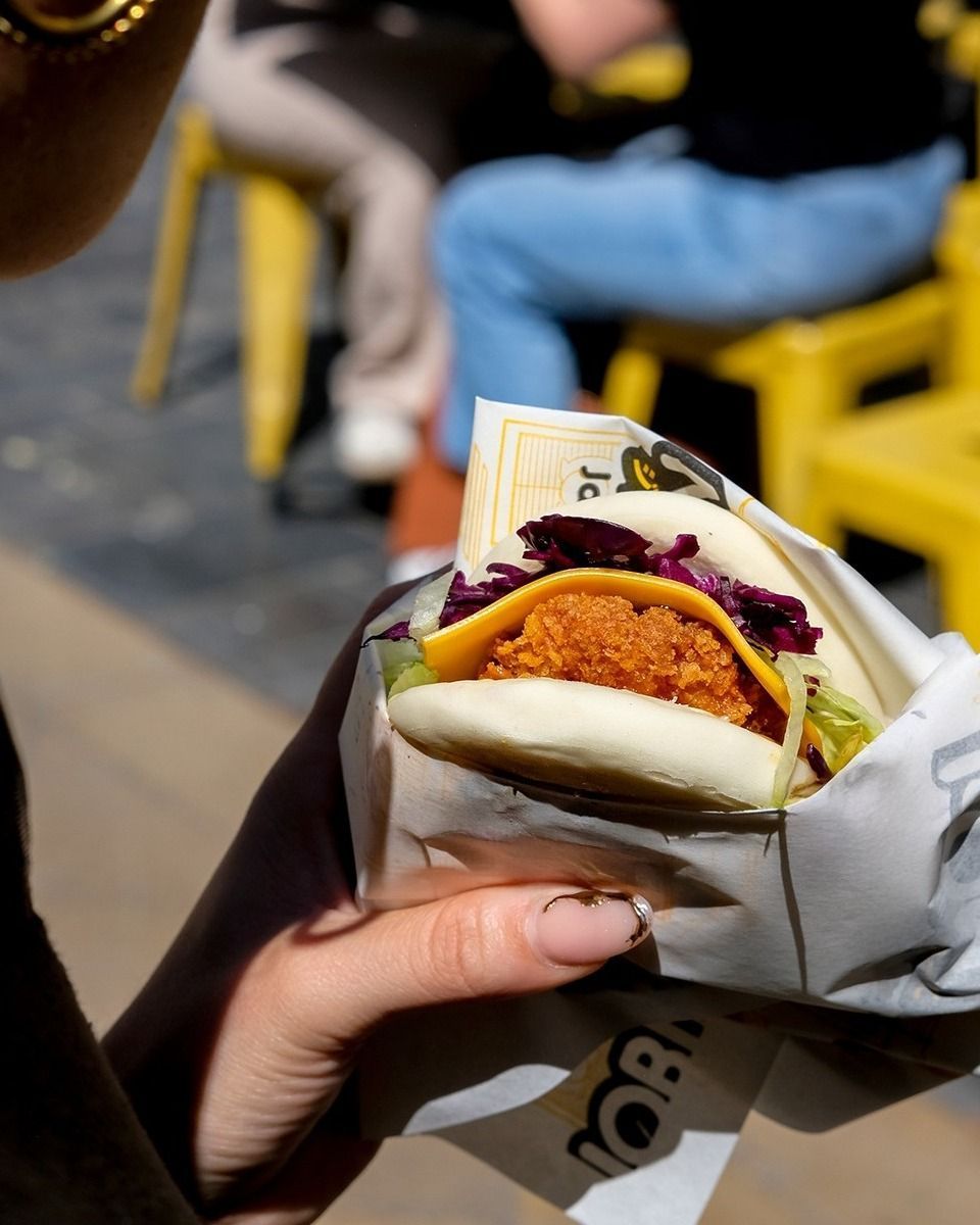 Hand holding a bao burger wrapped in paper with visible lettuce, cheddar cheese, purple cabbage, and a crispy fried patty.