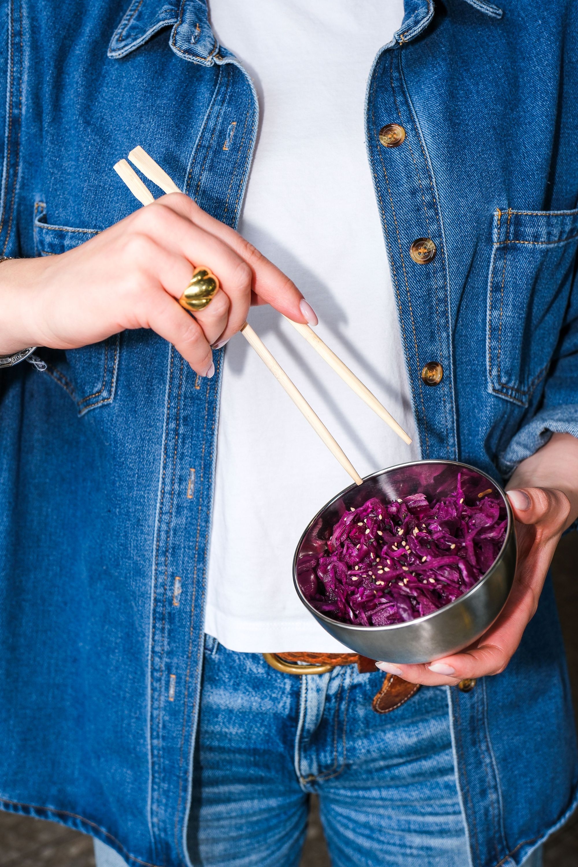 Person holding a bowl of purple cabbage salad with chopsticks wearing a denim jacket and jeans